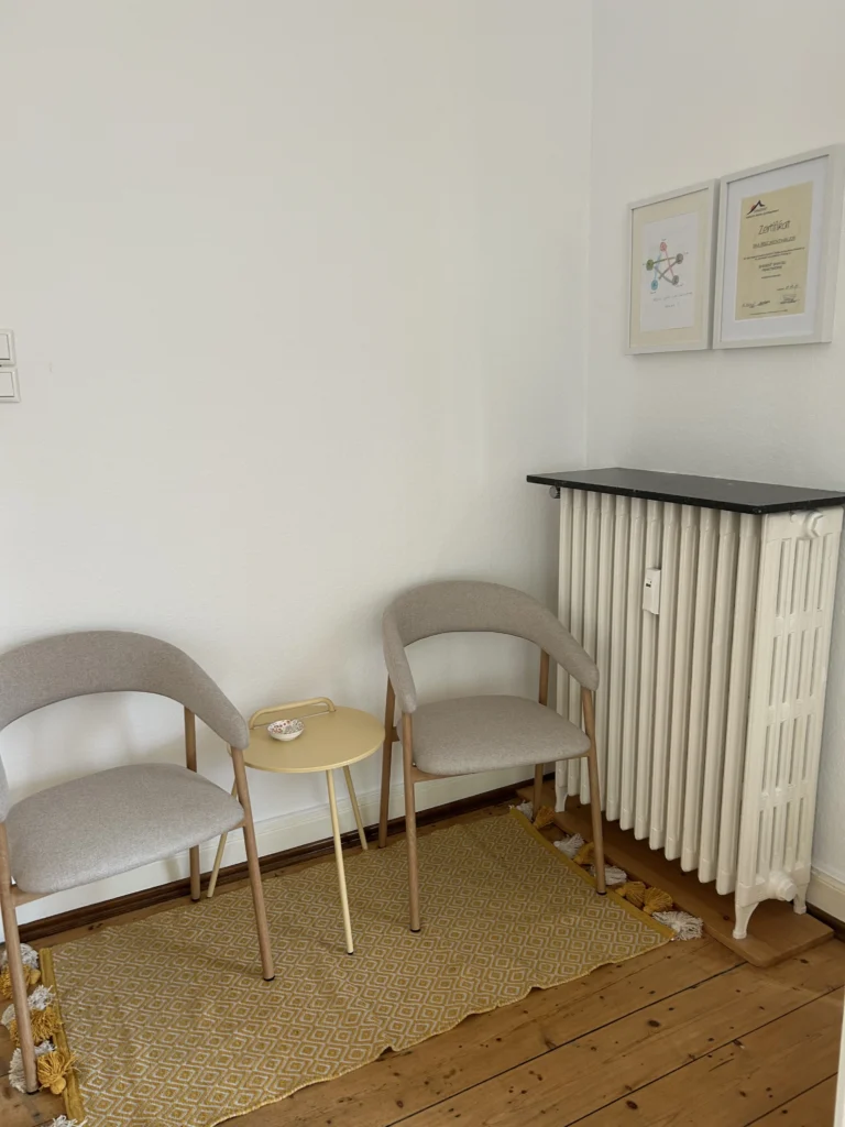 Shiatsu Krefeld Besprechungsbereich zeigt kleine Sitzecke mit zwei Stoffbespannten, St&uuml;hlen, kleinem gelbem Metalltisch und gewebtem Teppich mit gelb-wei&szlig; geometrischem Muster neben einer alten Heizung.